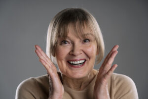 Full Mouth Dental Implants After Dentures: Your Options 1 Smiling older woman showing her healthy teeth with her hands raised beside her face.