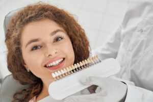 Porcelain Veneers vs Composite Veneers: Which Is Right for You? 2 Smiling woman comparing her teeth with a dental shade guide during a cosmetic dentistry consultation.