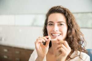 SureSmile vs Invisalign: Which Clear Aligner Is Right for You? 1 Smiling woman holding a clear orthodontic retainer in a dental clinic.